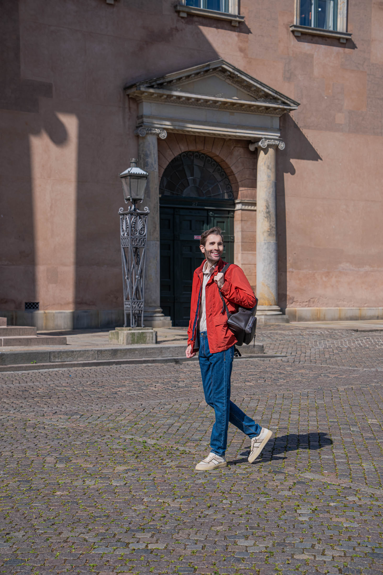 Marko Tadić walking past the dark green door and cast iron lantern of the Copenhagen Court House with a dark brown 1900 leather backpack on one shoulder