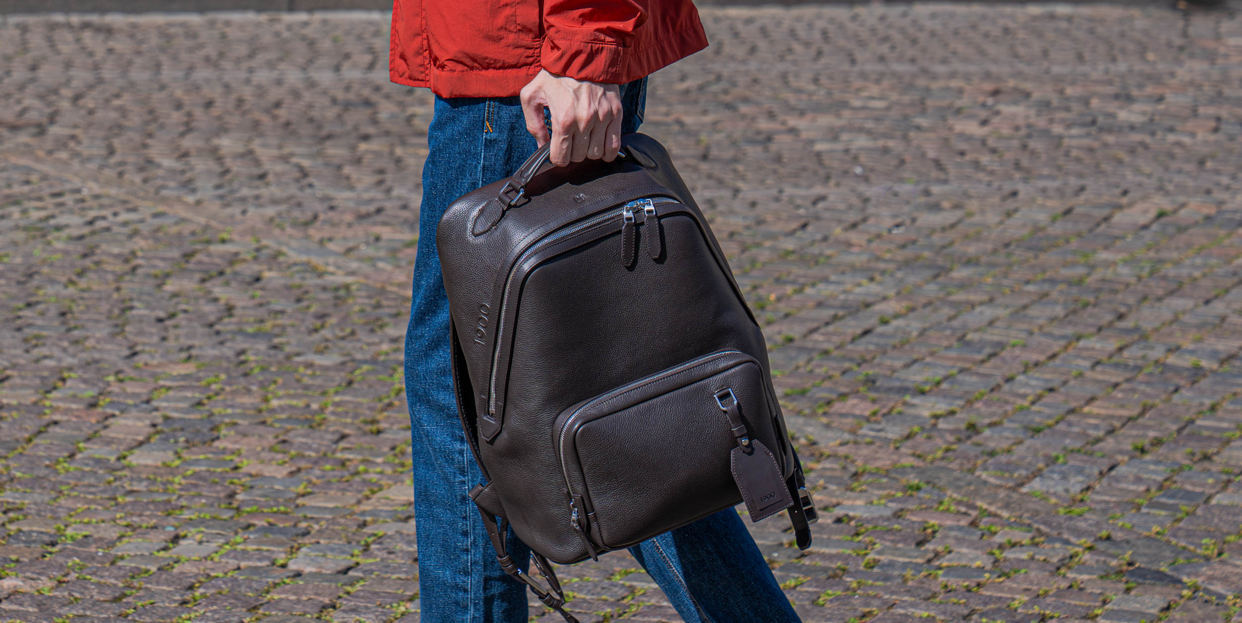 Marko Tadić holding a dark brown 1900 leather backpack by its top handle on Gammeltorv, Copenhagen, wearing a red Canali field jacket and Versace jeans
