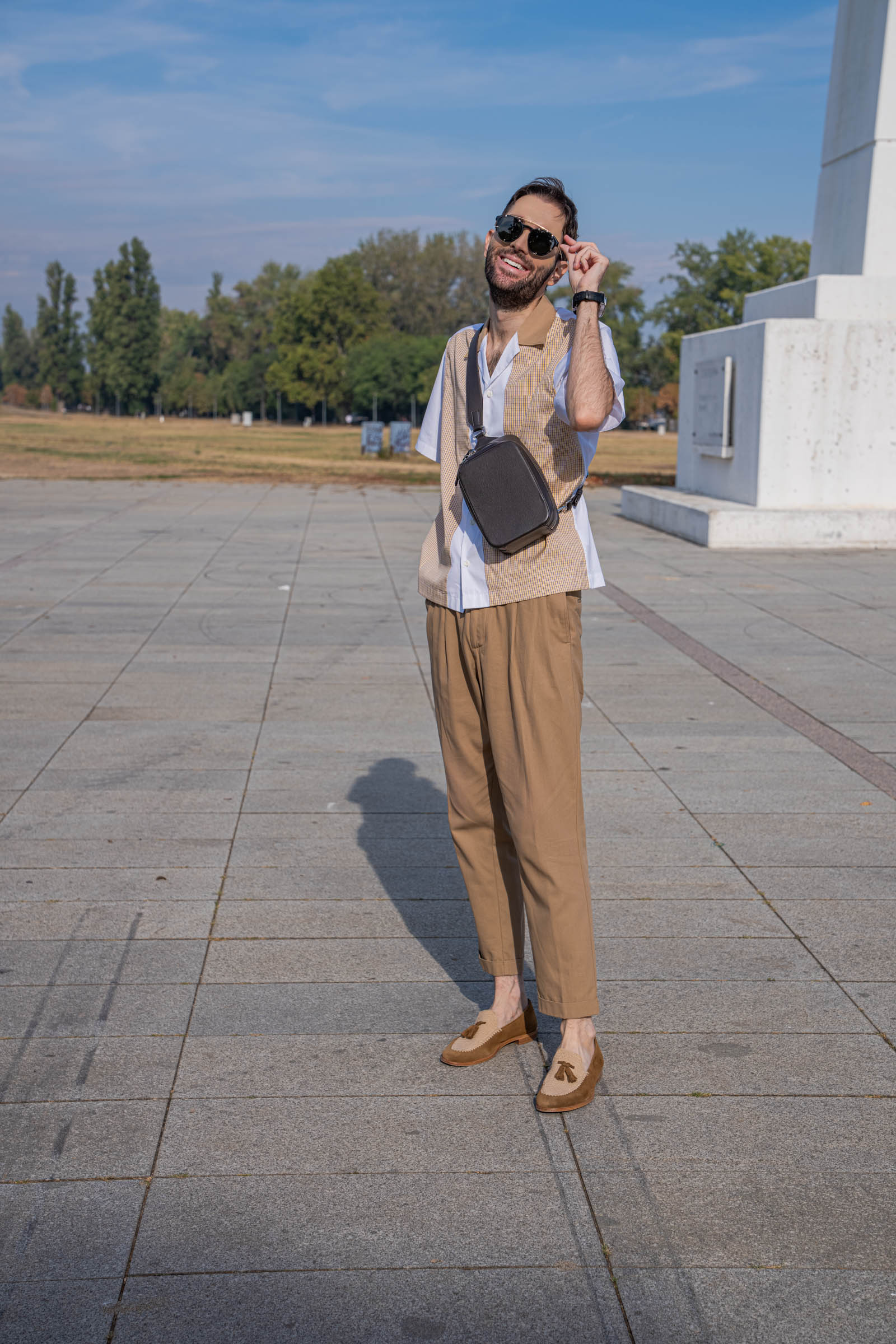 Marko Tadić adjusts his Givenchy round sunglasses while wearing a Dsquared2 paneled shirt, pleated trousers and the dark brown 1900 Tempo Grande Crossbody