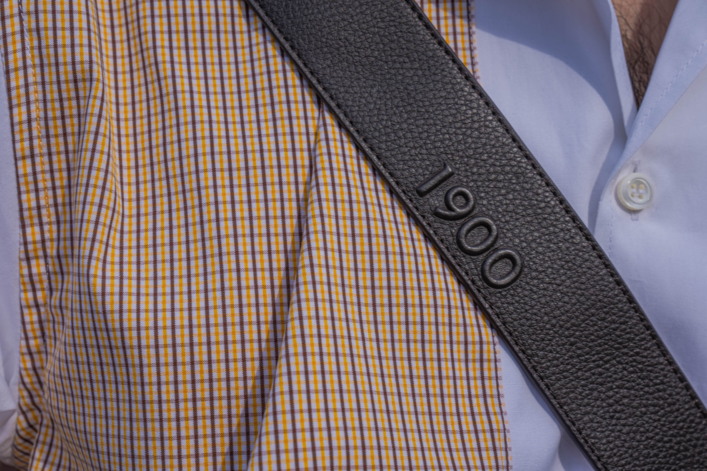 Close-up of the grained leather strap of the 1900 crossbody bag with embossed 1900 logo across the mustard and brown check of Marko Tadić's Dsquared2 shirt