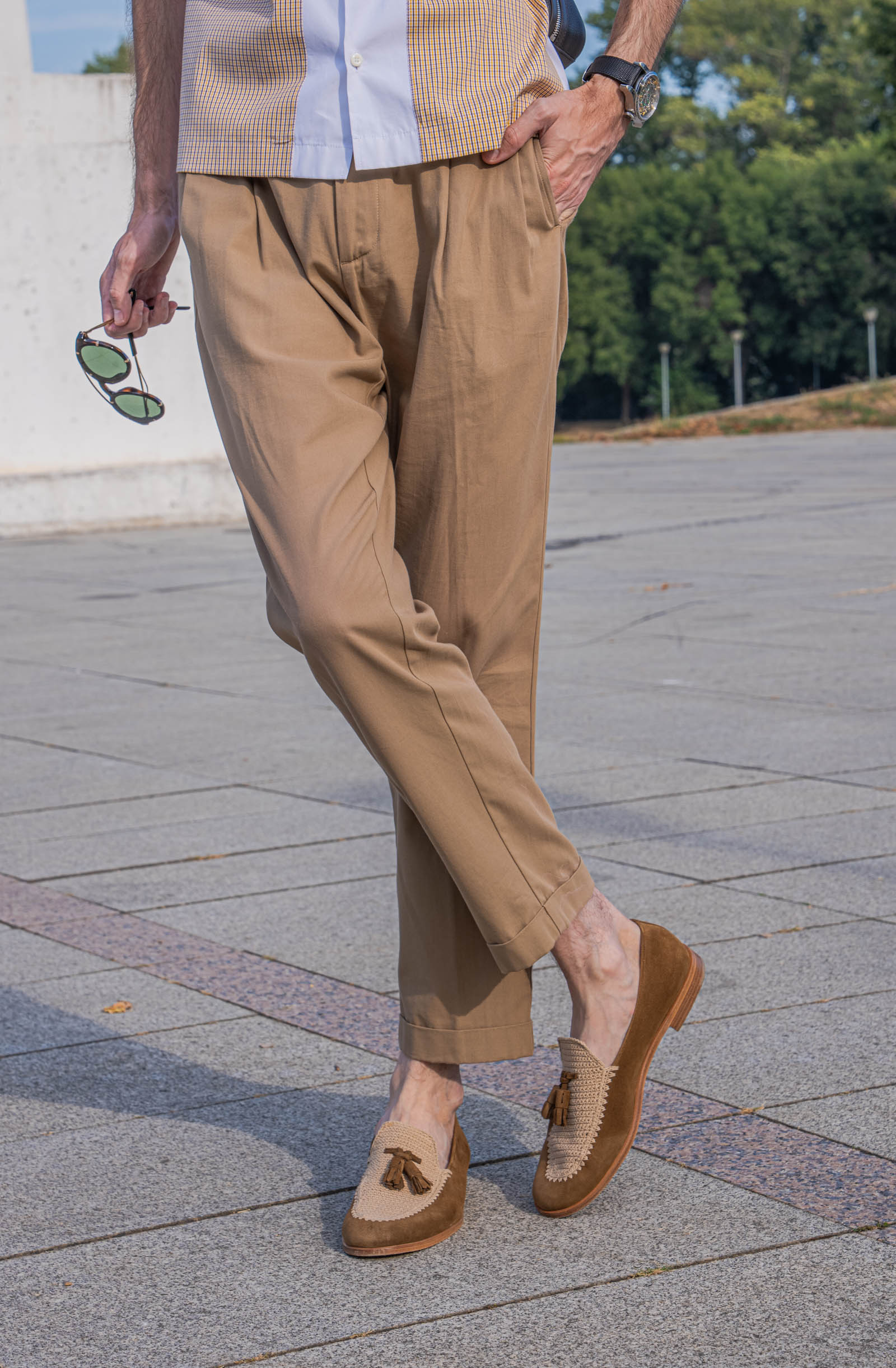 Crossed-leg pose by Marko Tadić highlighting a Dsquared2 outfit from the waist down: pleated tan trousers, green-lens Givenchy sunglasses and tassel loafers