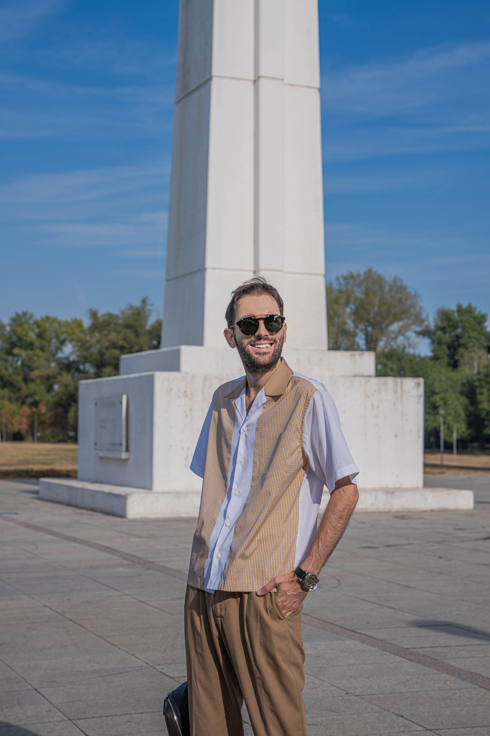 Smiling Marko Tadić in round Givenchy sunglasses and a Dsquared2 poplin shirt with mustard check panels, one hand in the pocket of his tan pleated trousers