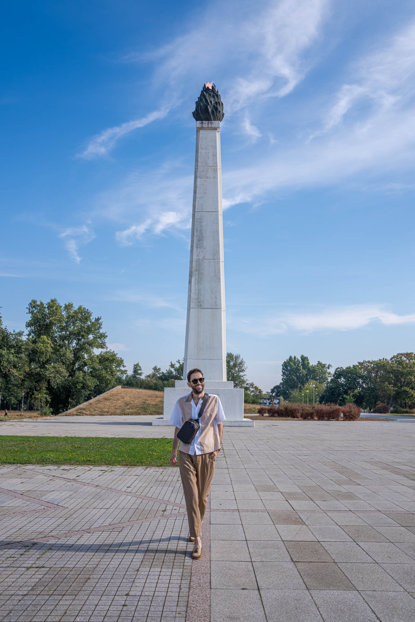 Marko Tadić walks toward the camera under a blue sky with light clouds, wearing a Dsquared2 outfit, the 1900 crossbody bag and Givenchy sunglasses