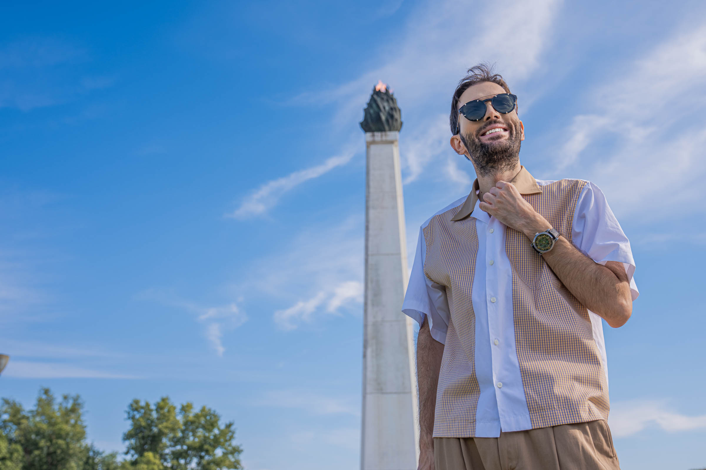 Low-angle portrait of Marko Tadić adjusting the camel collar of his Dsquared2 shirt, with Givenchy round sunglasses and the Bard's Whisper watch on his wrist