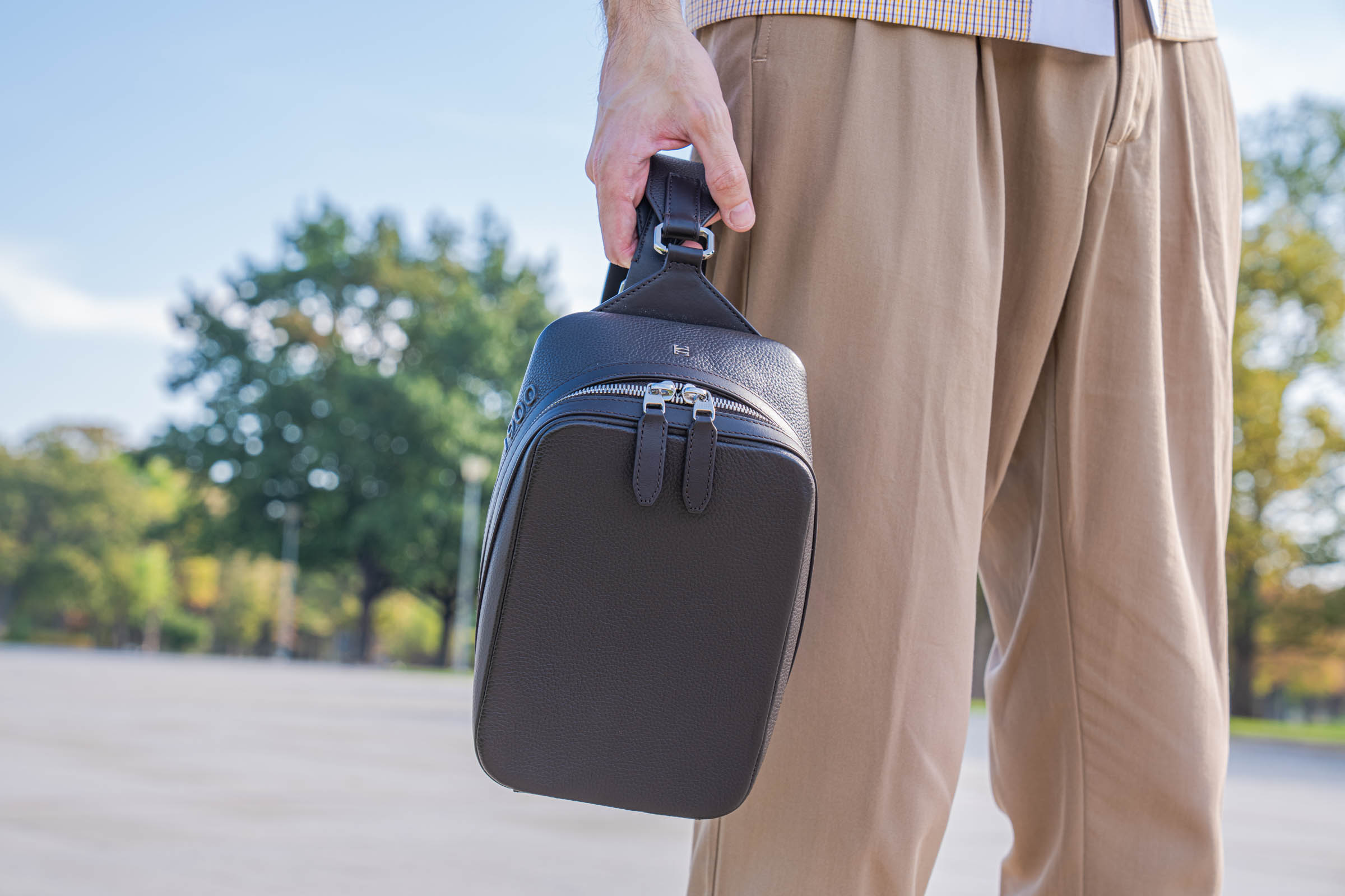Marko Tadić carries the dark brown 1900 Tempo Grande Crossbody bag by its strap loop, showing the octagonal metal ring and twin zips beside Dsquared2 trousers