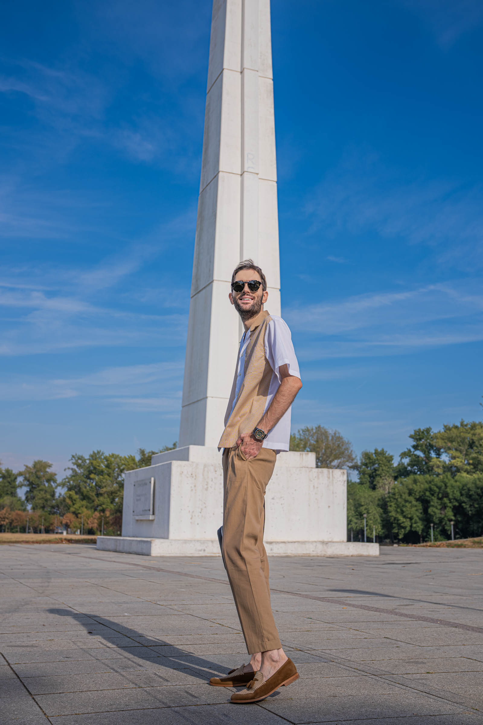 Side profile of Marko Tadić showing the relaxed drape of Dsquared2 pleated tan trousers with turn-ups, a white and check short-sleeve shirt and tobacco suede loafers