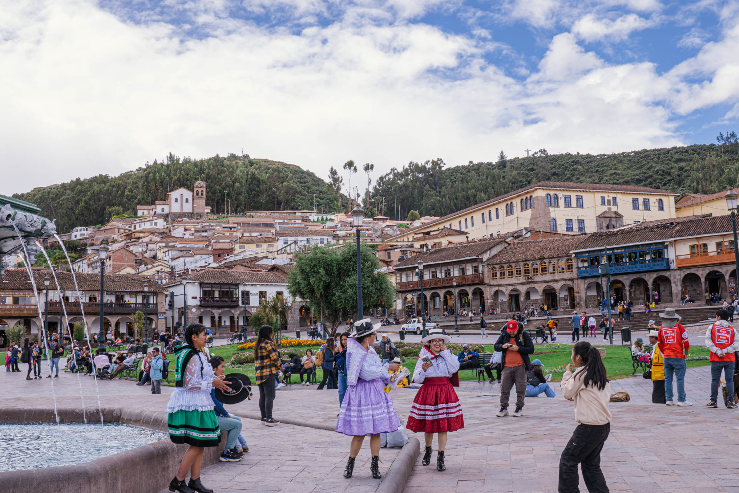 Panoramski pogled na Plaza de Armas u Kusku (Cuscu), Peru, sa zelenilom, kolonijalnim arkadama i brdovitim naseljem u pozadini.